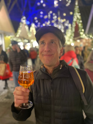 Mile holding a glass of amber drink at a festive, illuminated market.